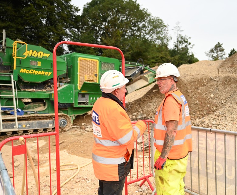 Two demolition workers discussing workload in an exclusion on a demolition site, next to a crusher and screener.